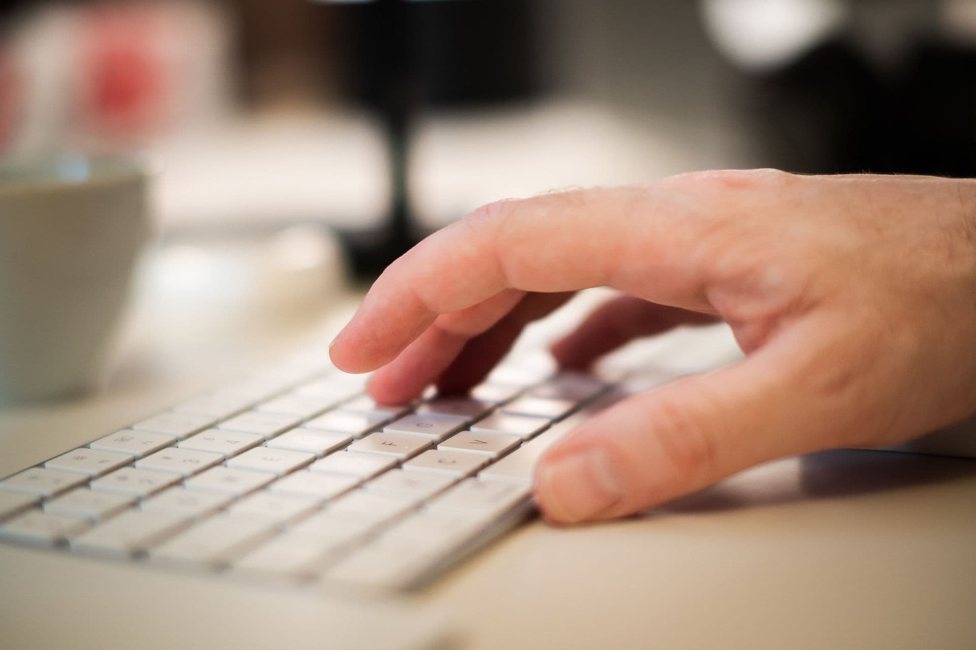 Nahaufnahme einer Hand auf einer hellen Tastatur, im Hintergrund eine Kaffeetasse, ruhige Lichtstimmung